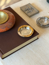 Hermes horseshoe ashtray on a brown book beside a red stone ashtray, a silver coaster, and a pewter box on travertine