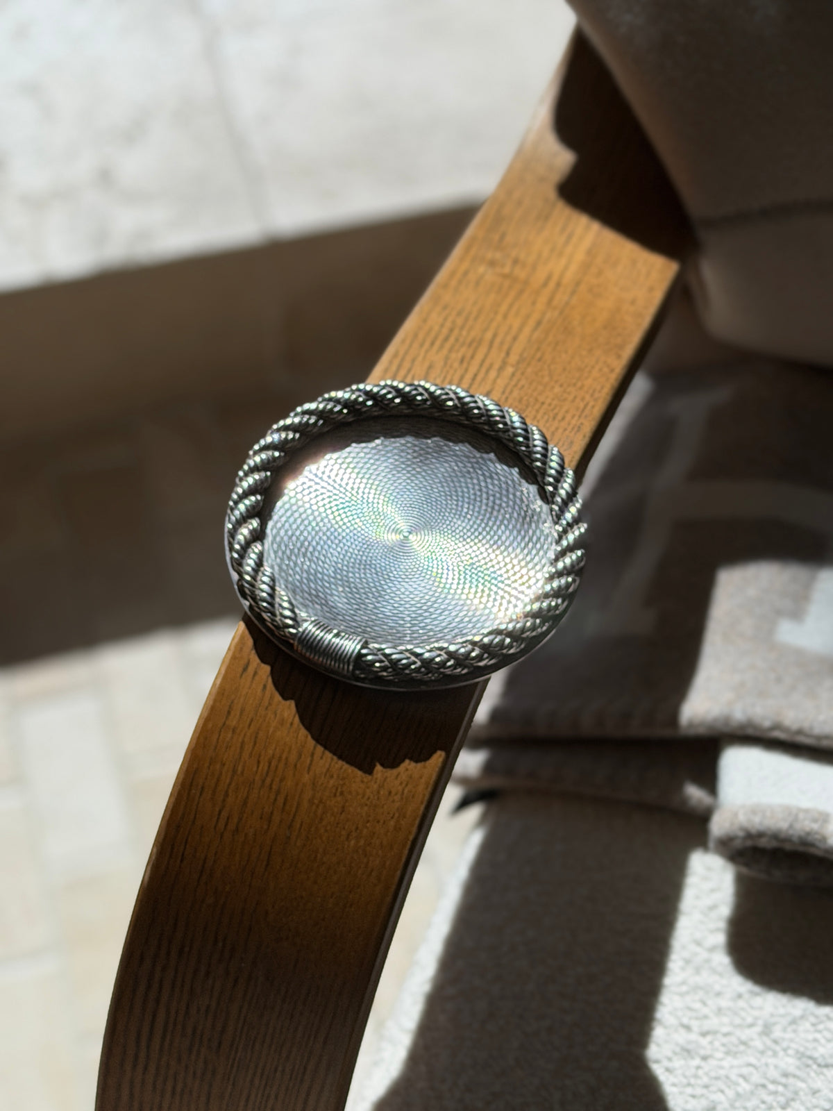 Hermes guilloche coaster balanced on a bentwood oak chair arm in sunlight, the engine turning shimmering with iridescent light