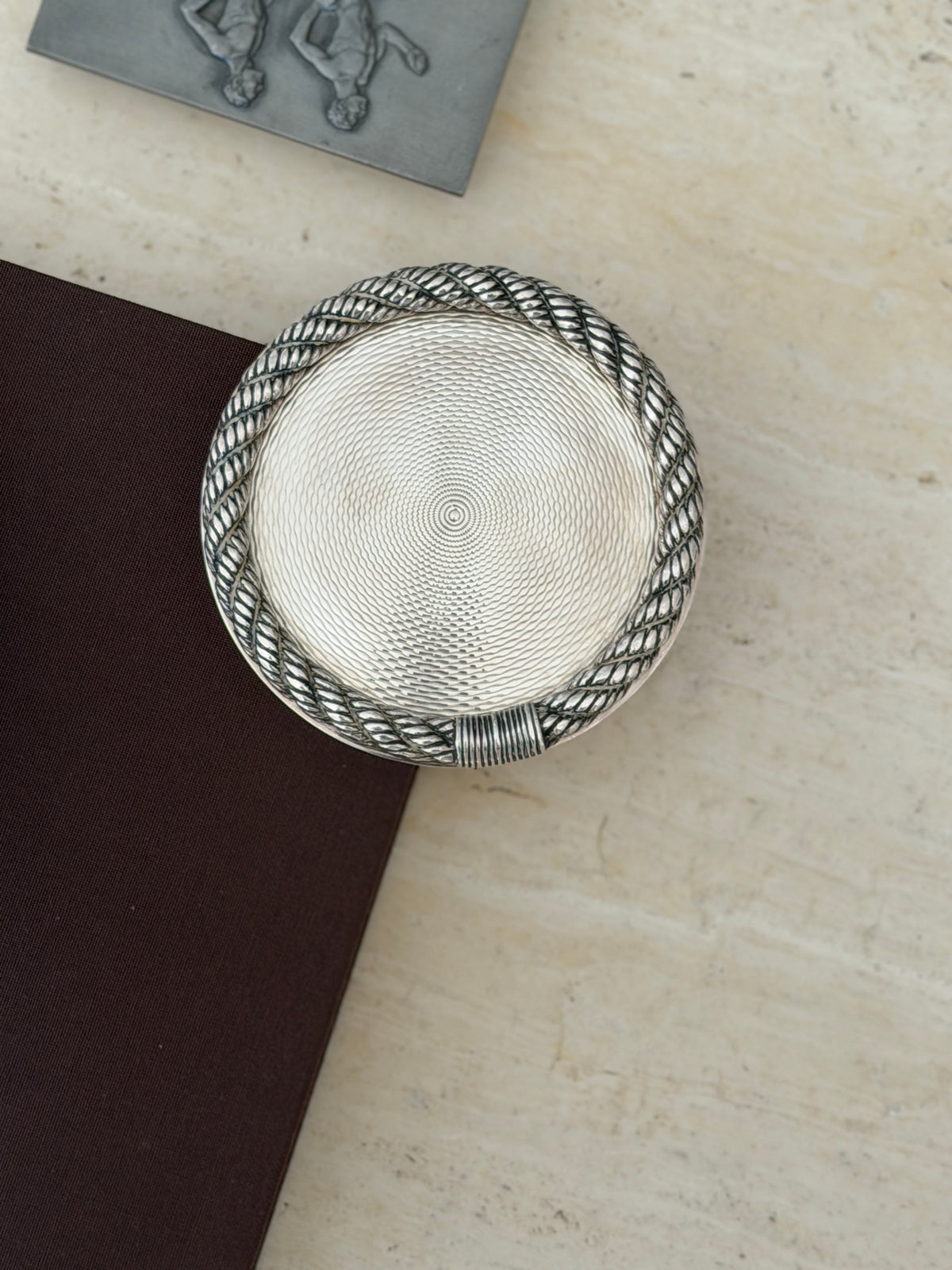 Top down view of the Hermes rope rim coaster on the corner of a brown book beside a pewter relief box
