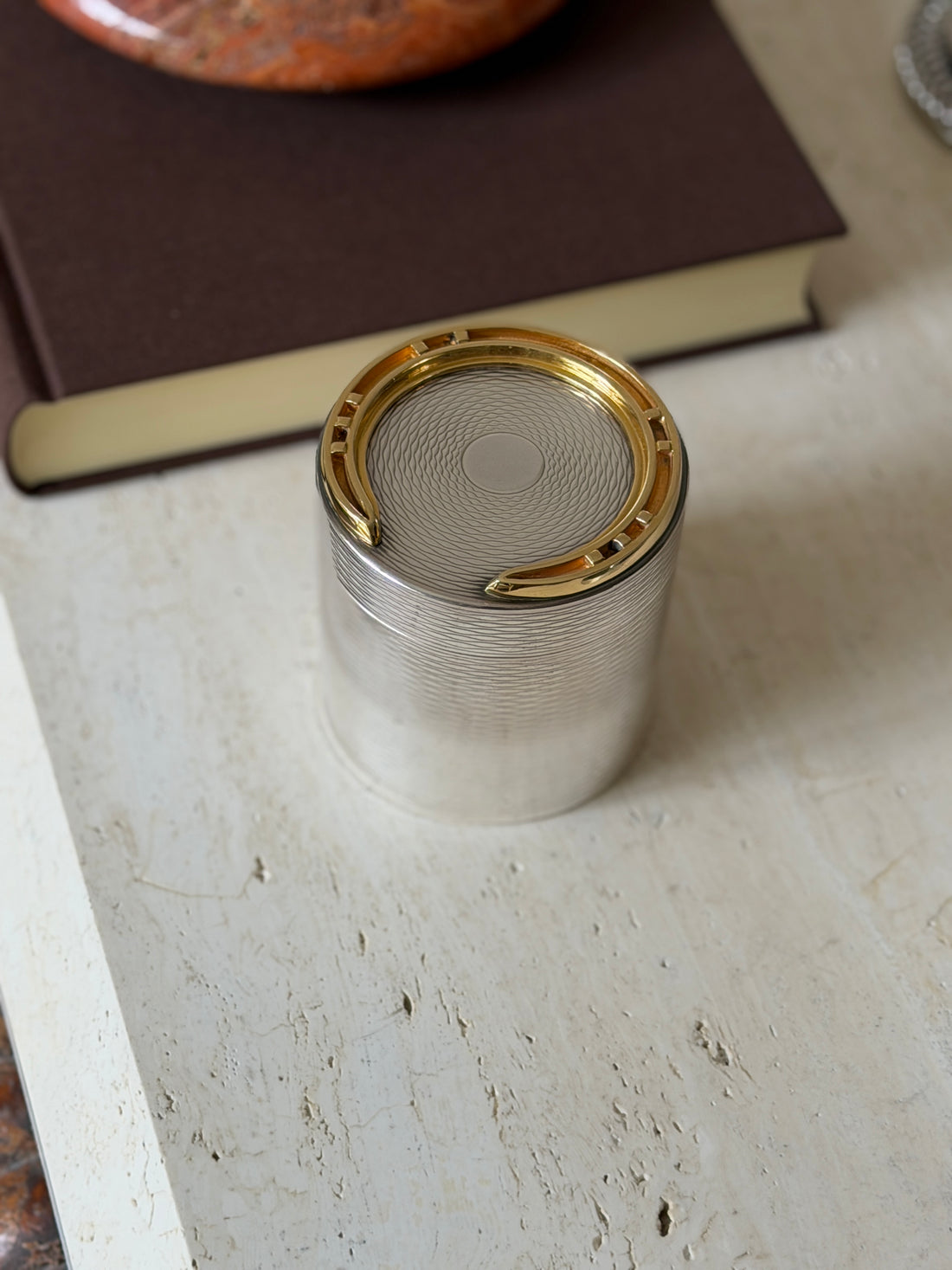 Hermes cylindrical cigarette box with engine-turned silver-tone body and a gilt brass horseshoe applied to the guilloche lid, beside a brown book on travertine