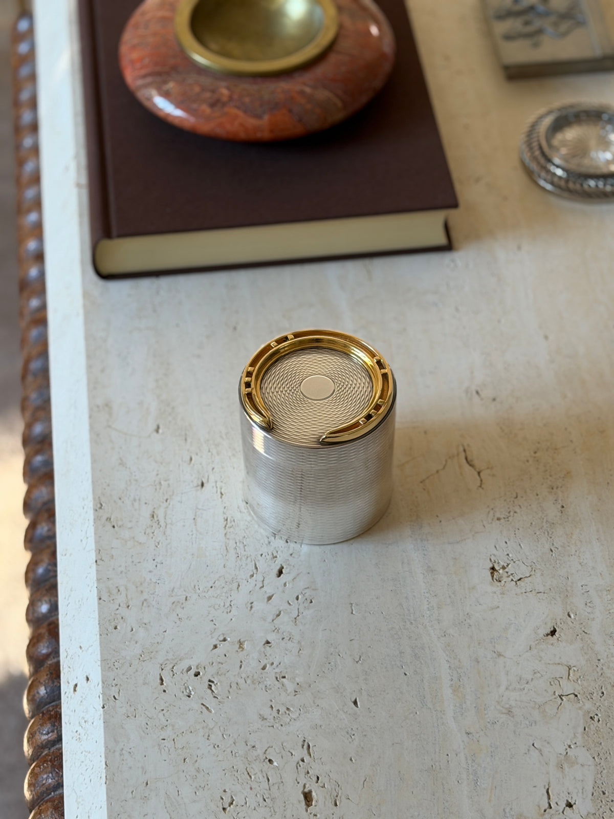 Hermes horseshoe cigarette box on a travertine table with a red stone ashtray on a brown book and a silver coaster behind