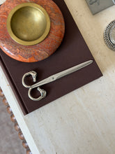 Overhead view of the closed Hermes scissors with horse-head handle rings resting on a brown book, next to a stone bowl with a brass rim