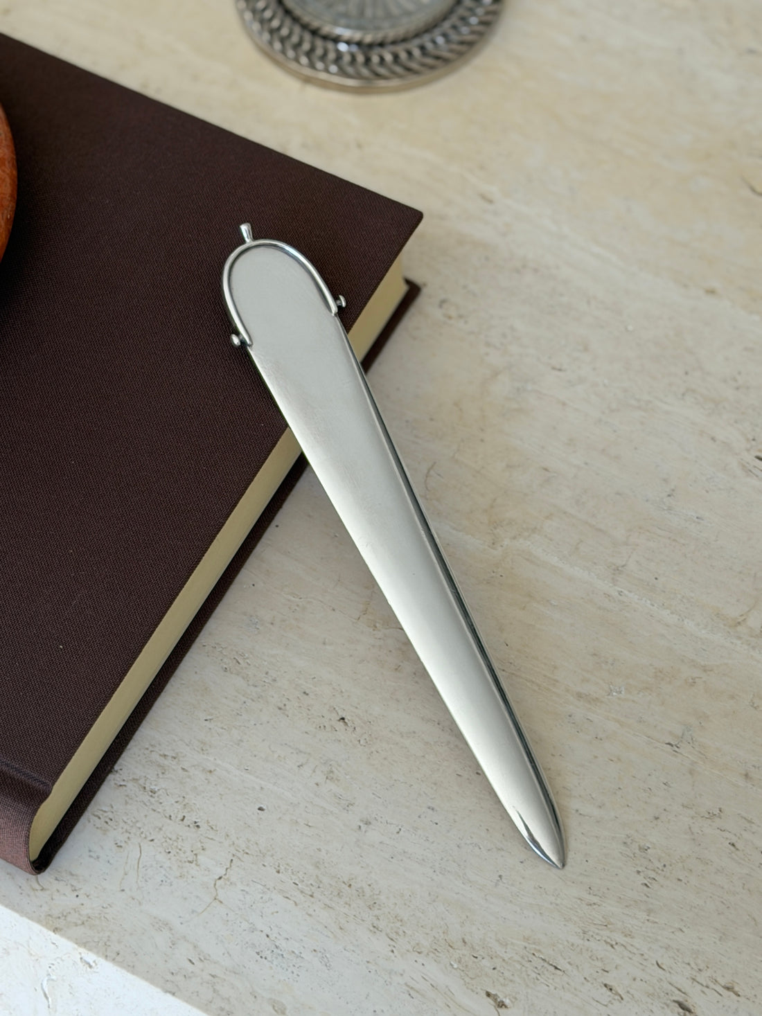 Hermes spur handle letter opener angled against the corner of a brown book, light catching the smooth rounded handle and blade edge
