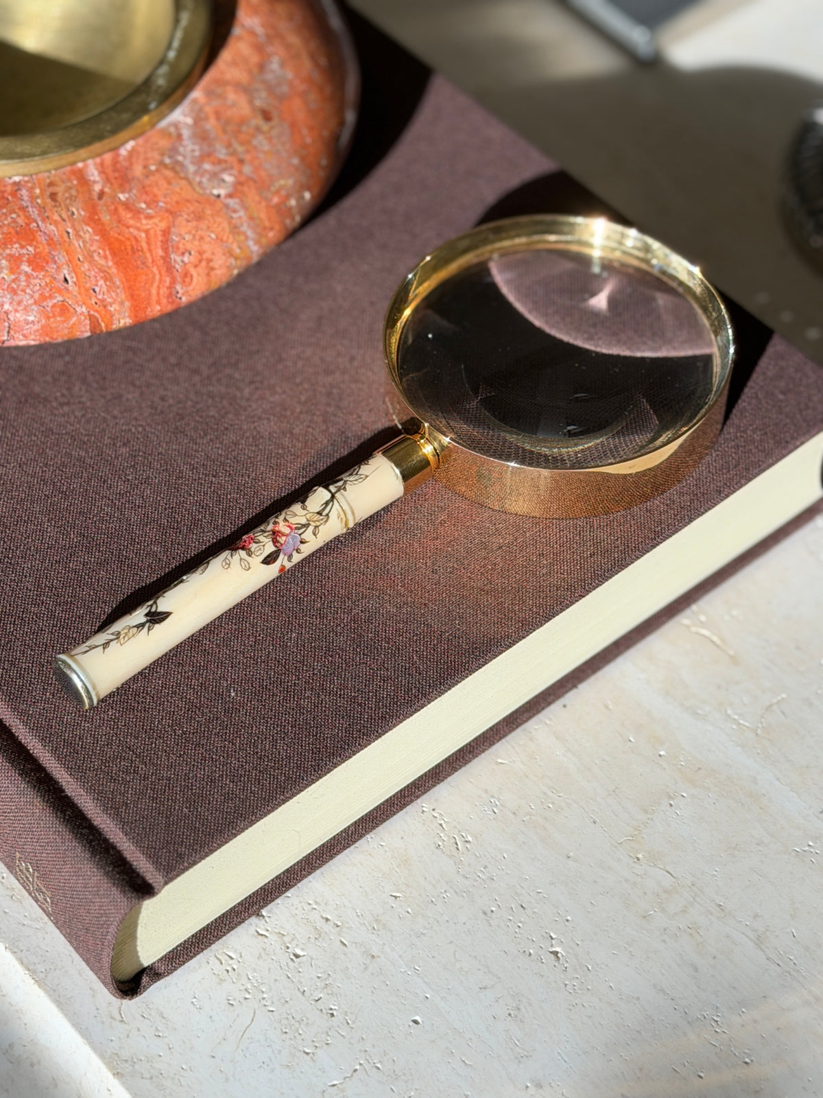 Hermes magnifying glass in afternoon light on a brown book, the inlaid lavender and red flowers glowing along the handle