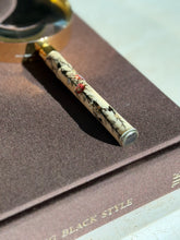 Close up of the handle end on a brown book, the silver cap and inlaid red blossoms, leaves, and a beetle along the cream handle