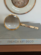 Front view of the Hermes magnifying glass standing on a stack of books, a gilt rimmed porcelain plate in the background