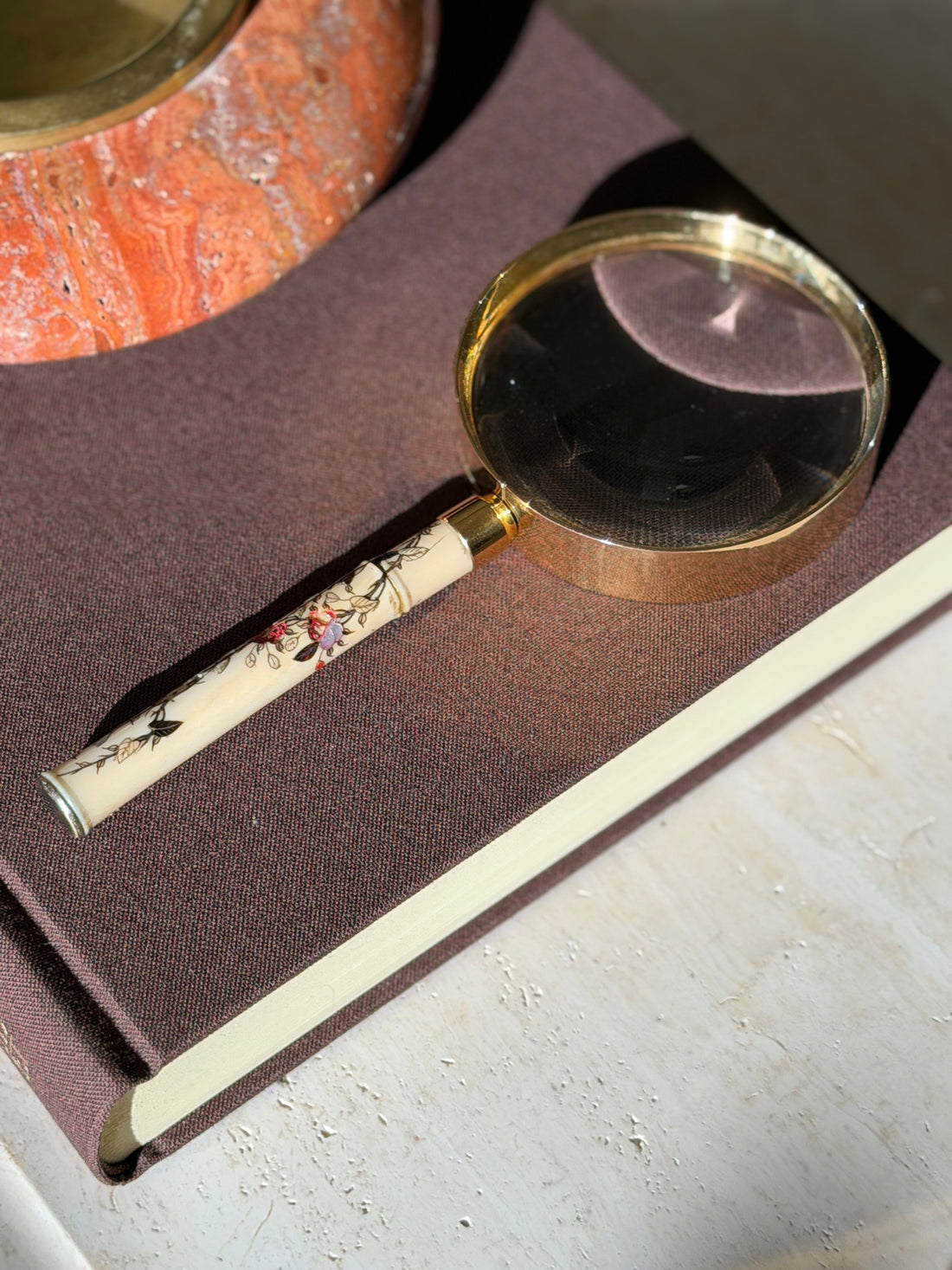 Hermes magnifying glass on a brown clothbound book in strong sunlight, beside a red travertine ashtray with a brass insert