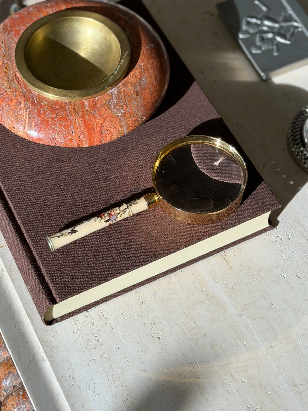 Overhead view of the Hermes magnifier on a brown book beneath a red stone and brass ashtray, the lens magnifying the linen weave