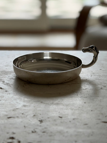 Hermes silver plated catchall seen at eye level on travertine, a round shallow dish with a sculpted horse head handle rising from the rim