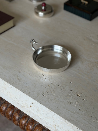 Round silver plated dish near the travertine table edge and its bobbin turned wood apron, a lidded box and dark box blurred behind