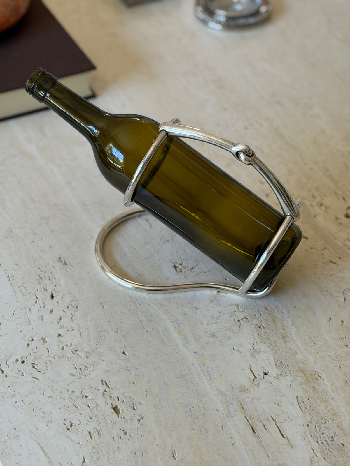 Hermes horse bit wine caddy holding an olive green bottle at a pouring angle on a travertine table