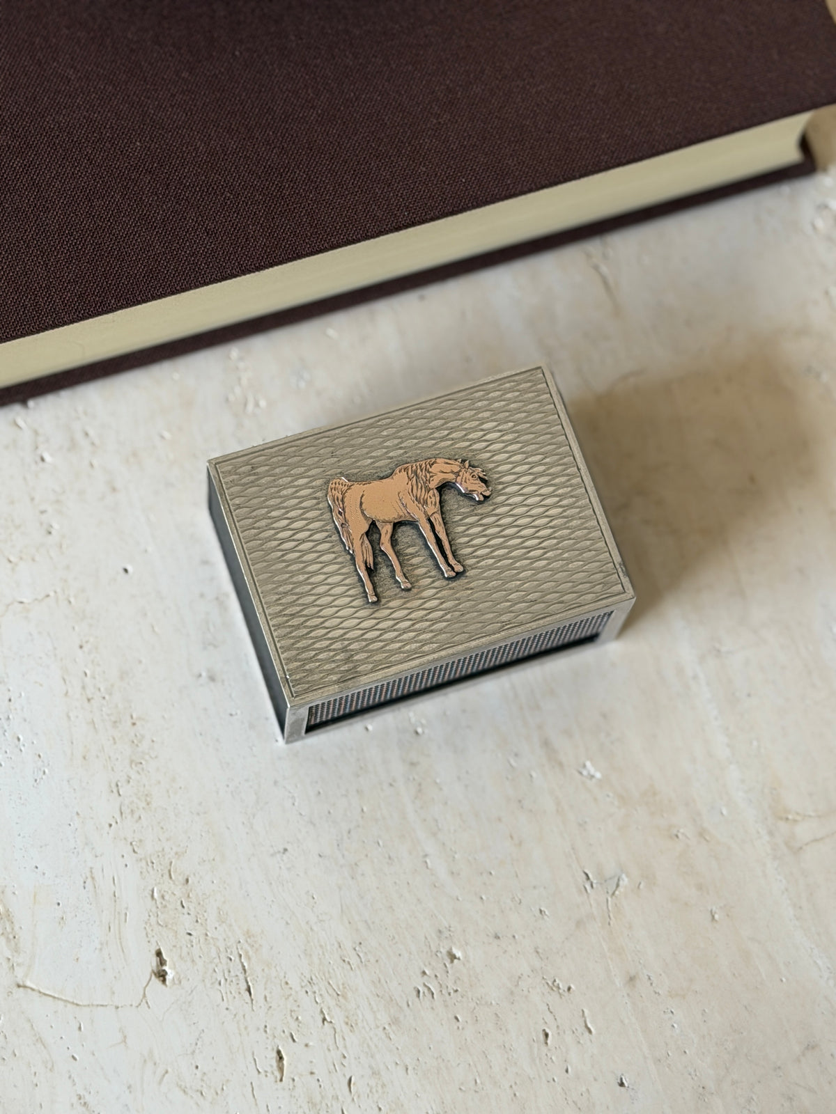 Hermes solid silver matchbox cover with an applied gilt horse on an engine turned wave pattern lid, resting on travertine beside a brown book