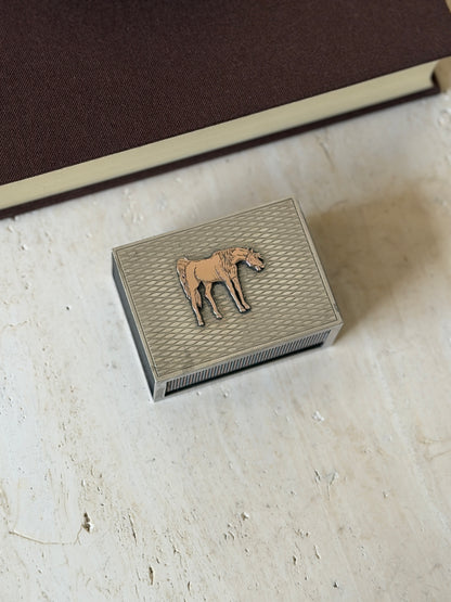 Hermes solid silver matchbox cover with an applied gilt horse on an engine turned wave pattern lid, resting on travertine beside a brown book