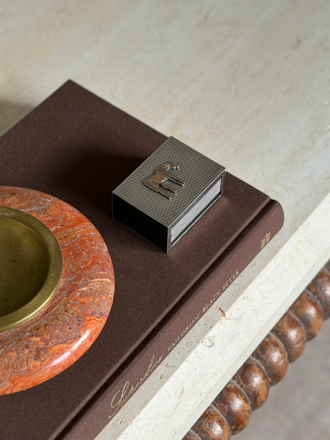 Hermes silver and gilt horse matchbox on a brown book beside a red stone ashtray with brass insert on a travertine console