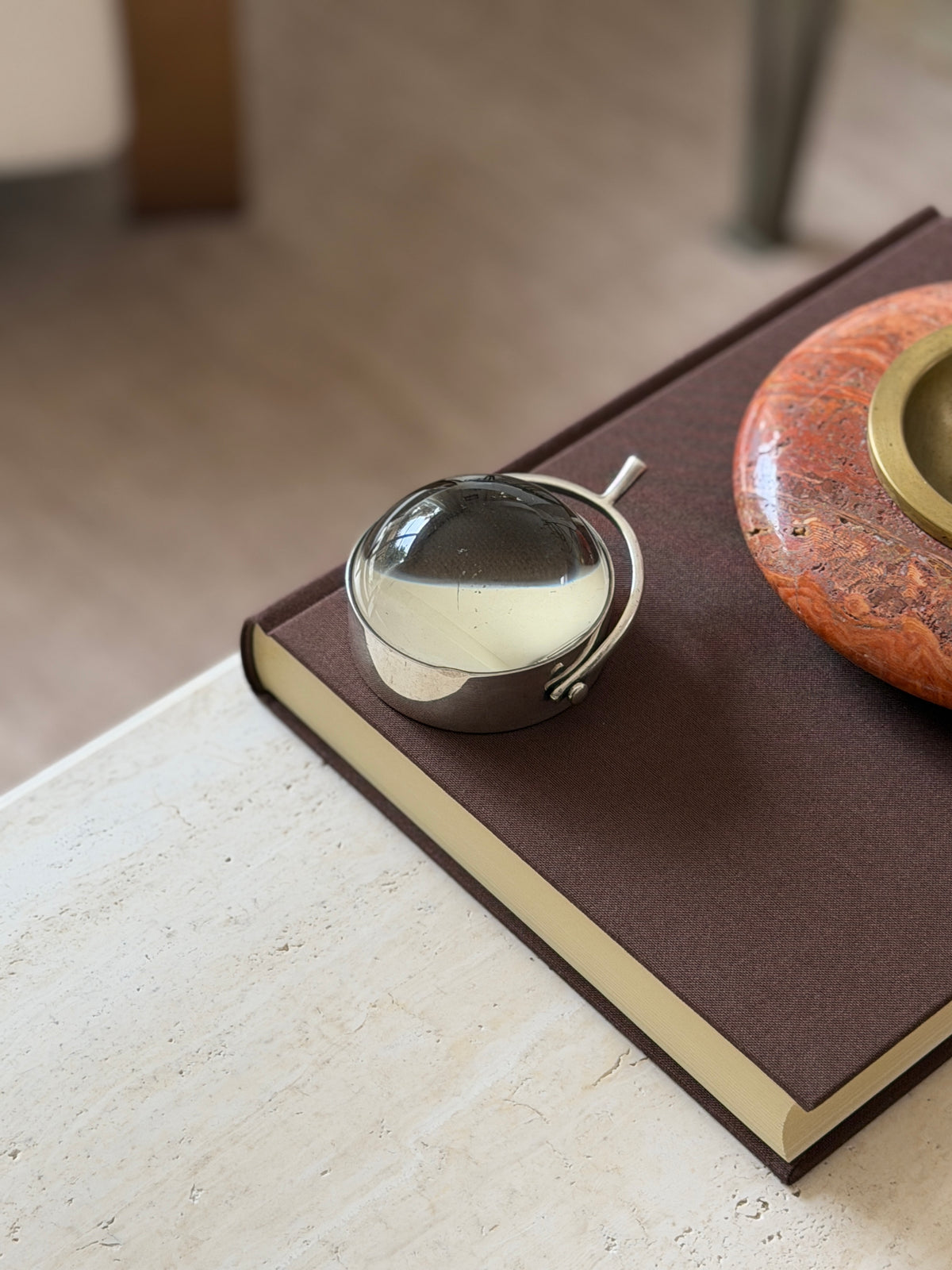 The round magnifying glass with its domed lens and spur-shaped handle resting on a book, an orange stone bowl blurred in the background