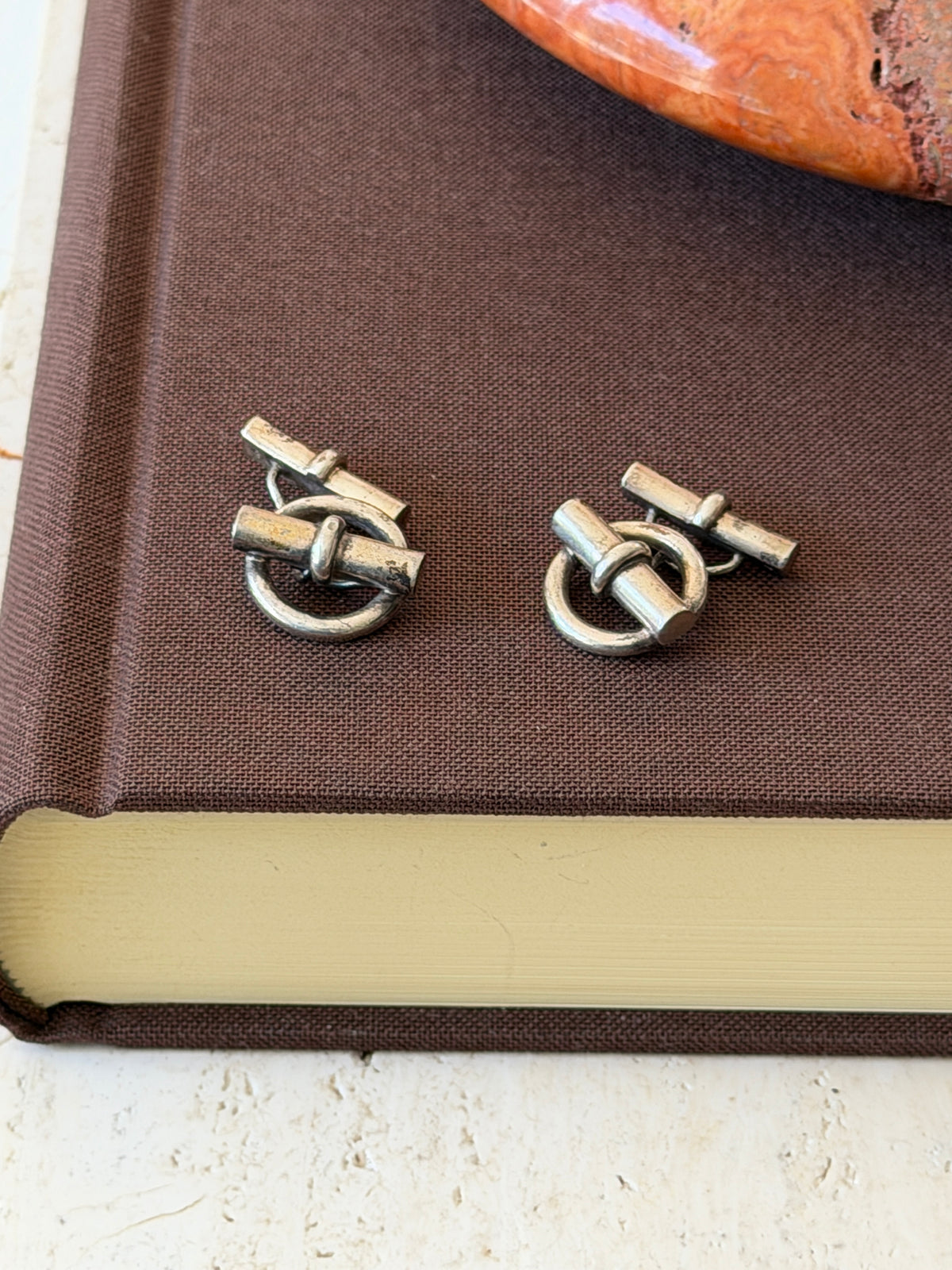 Pair of silver toggle cufflinks with ring fronts crossed by banded bars, resting on a brown clothbound book beside a red stone dish
