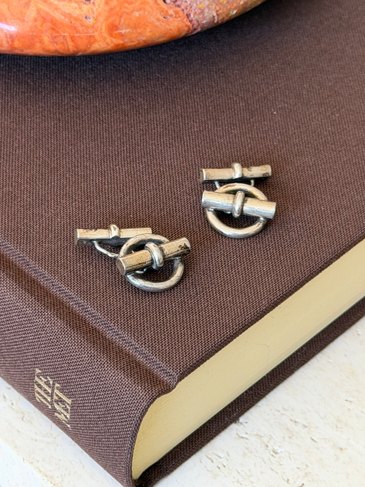 Angled view of the ring and toggle cufflinks on the corner of a brown book, patina visible on the rounded bars