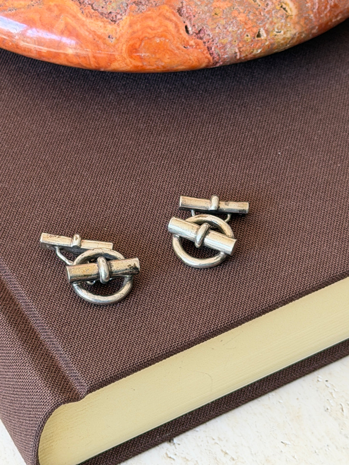 Two ring and bar cufflinks side by side on a brown book cover in soft daylight, a banded stone dish above