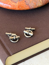 Two ring and bar cufflinks side by side on a brown book cover in soft daylight, a banded stone dish above