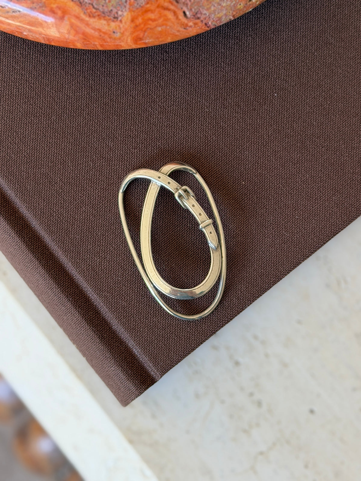 Hermes sterling silver money clip formed as an oval loop wrapped by a belt strap with buckle, keeper, and pierced holes, on a brown book