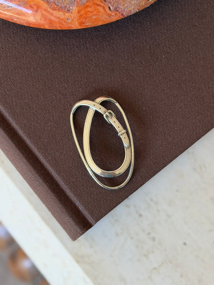 Hermes sterling silver money clip formed as an oval loop wrapped by a belt strap with buckle, keeper, and pierced holes, on a brown book