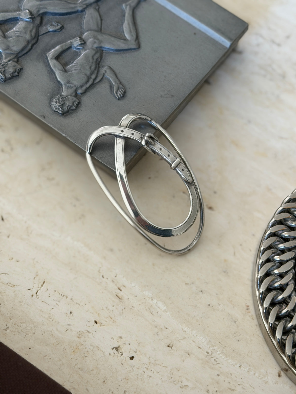 Belt strap money clip leaning against a pewter relief box on travertine, showing the sprung double loop