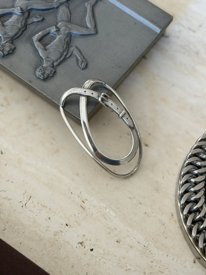 Belt strap money clip leaning against a pewter relief box on travertine, showing the sprung double loop