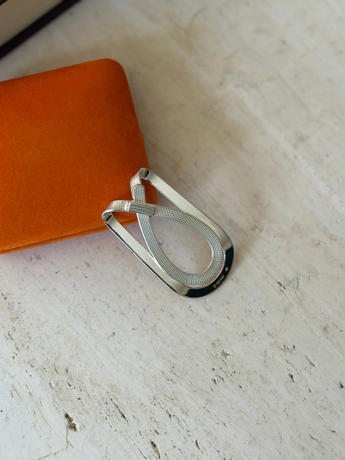Hermes crossed strap money clip resting against the orange suede box, sun catching the textured strap and polished silver