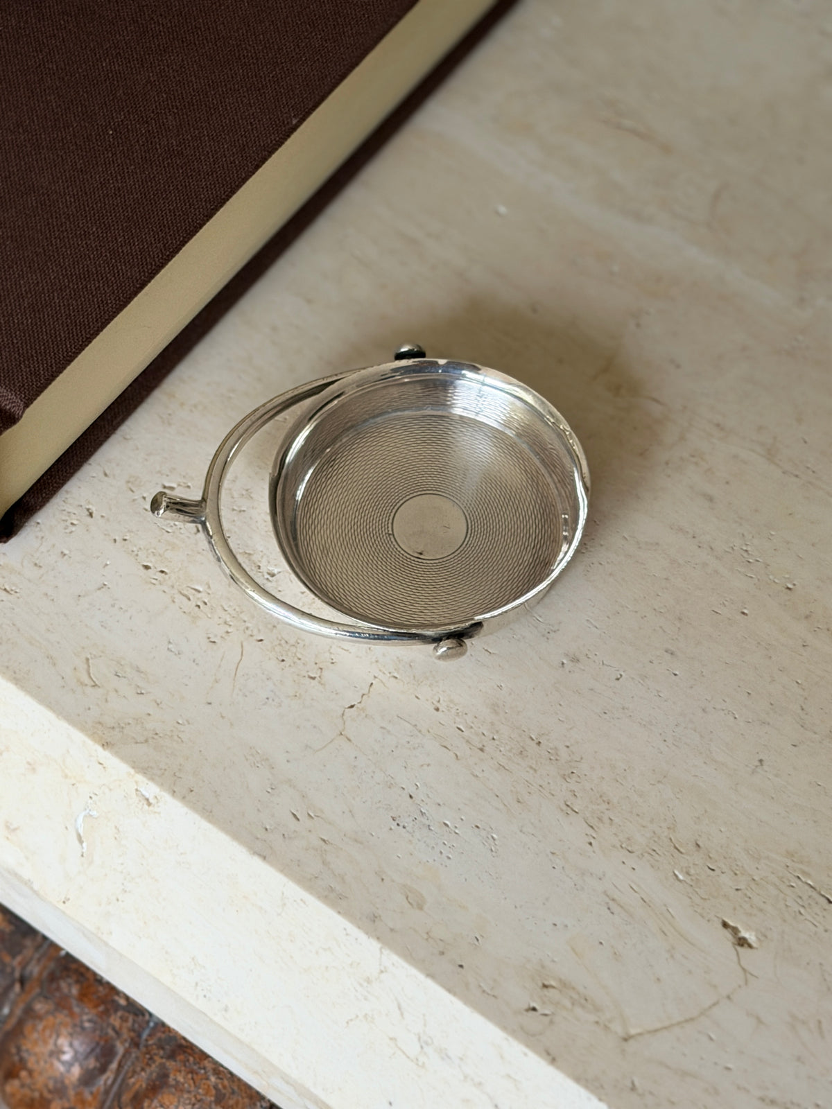 Sterling spur catchall on travertine near a brown book, the hoop handle pivoting on ball ends with a short projecting stem like a spur neck