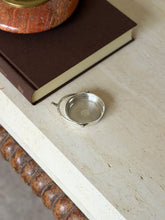Small sterling silver spur ashtray on a travertine table beside a brown book topped with a red stone ashtray
