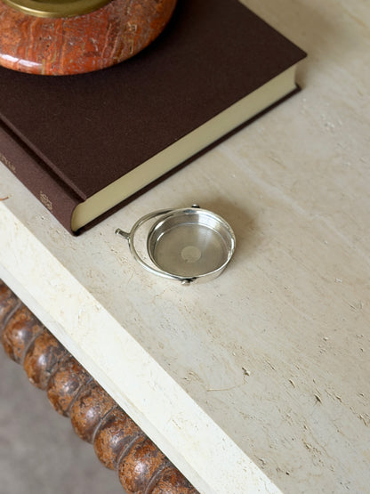 Small sterling silver spur ashtray on a travertine table beside a brown book topped with a red stone ashtray
