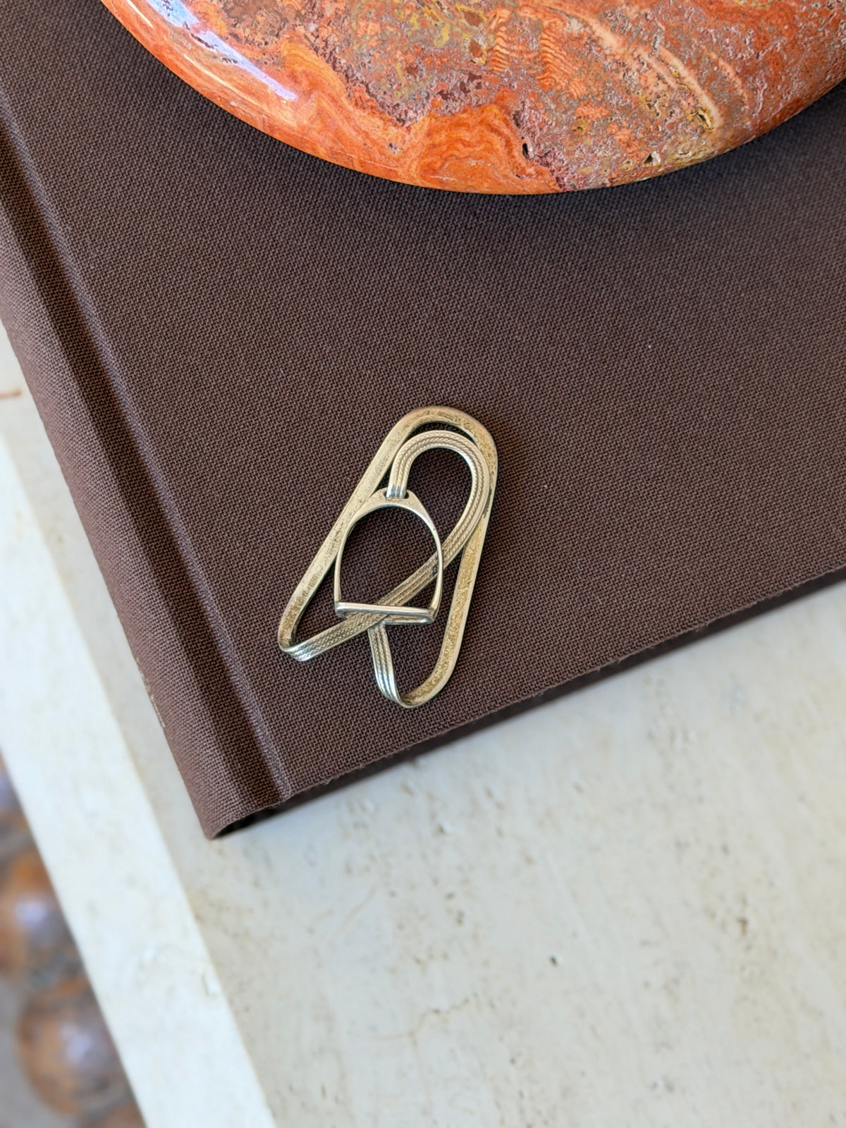 Hermes stirrup money clip on a brown book, a miniature stirrup threaded on an engraved strap inside an oval silver loop