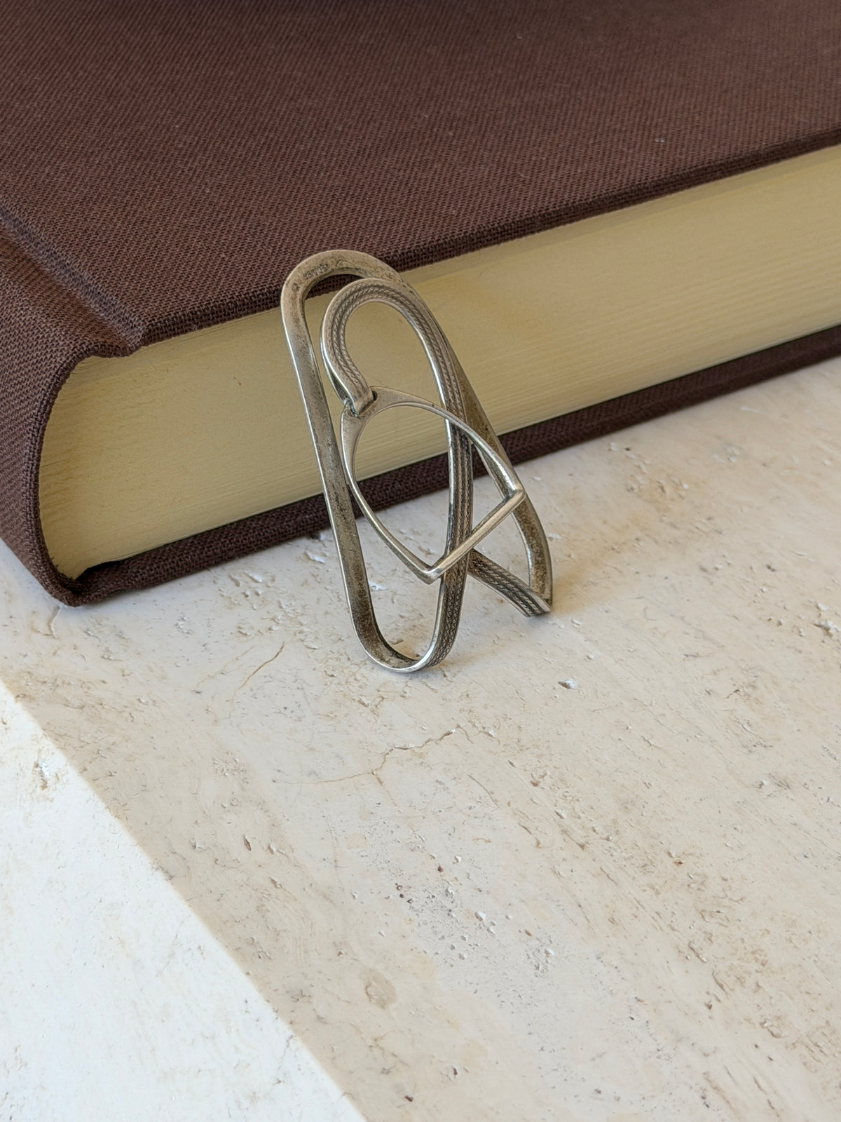 Stirrup money clip standing against a brown book on travertine, the worn patina showing along the plain outer loop