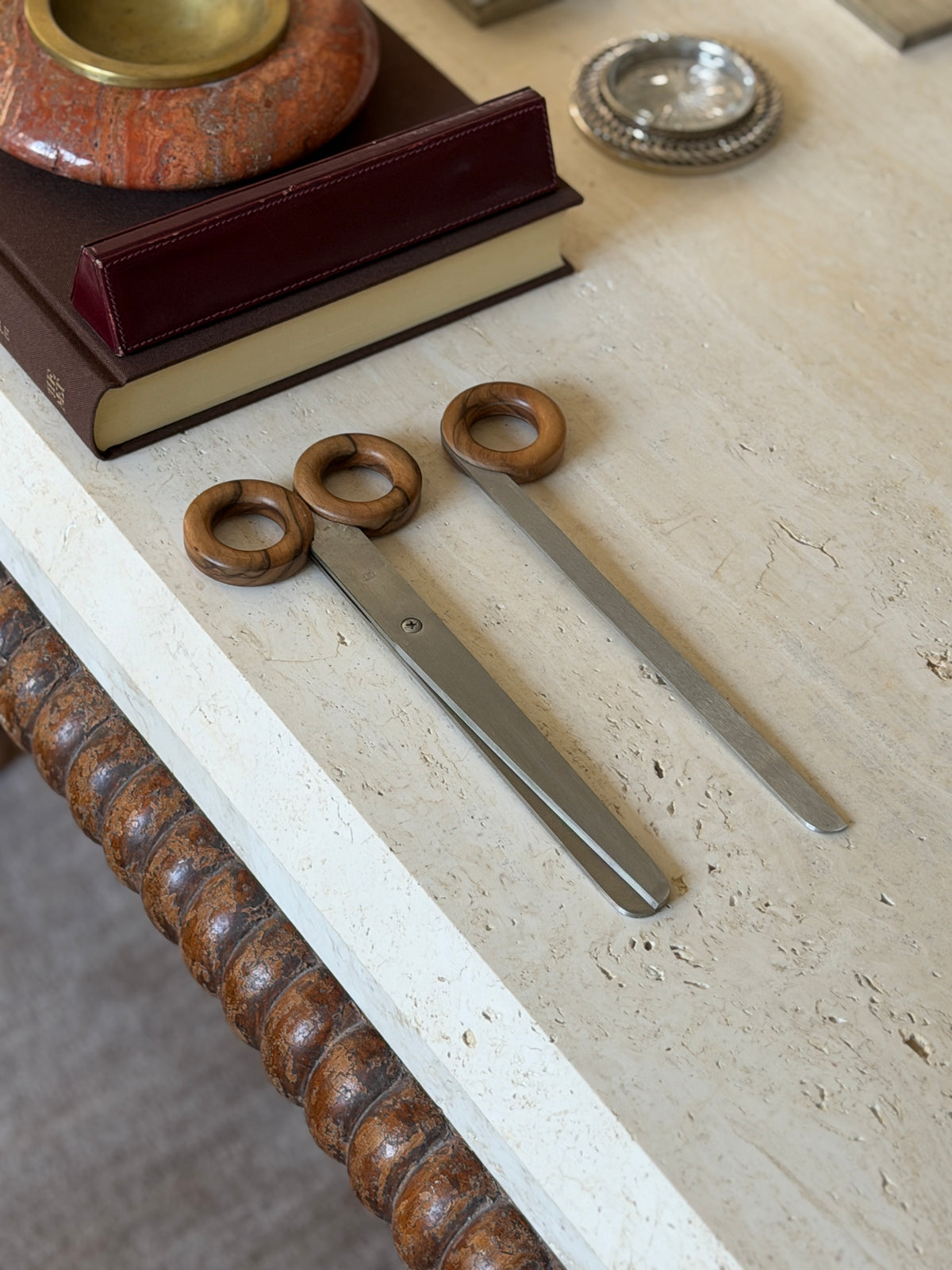 Scissors and uncased letter opener arranged on a travertine table, with the oxblood leather case resting on a brown book behind them