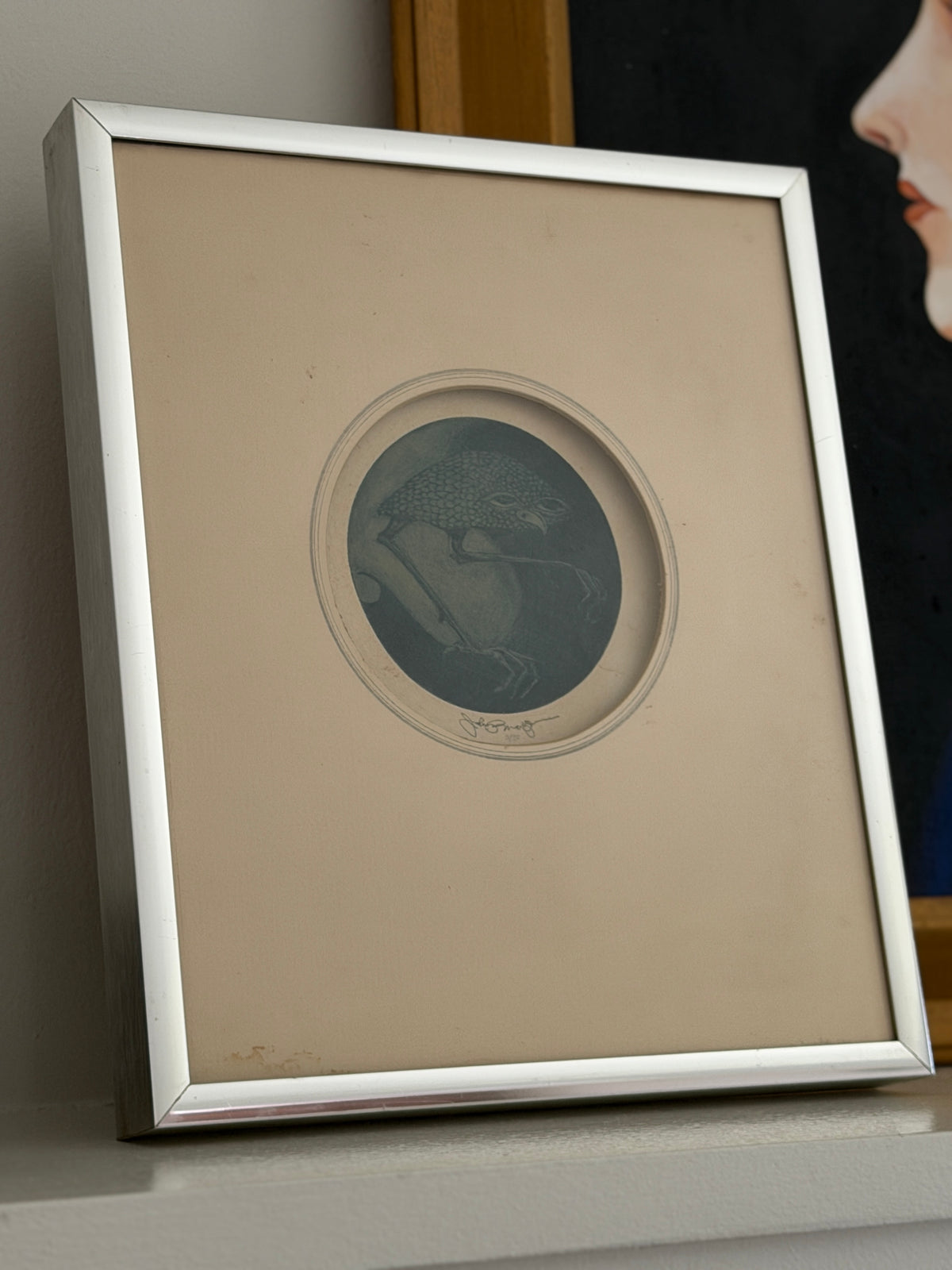 Angled view of the framed John P. Morgan print on a shelf, the silver frame edge catching light against a dark portrait behind