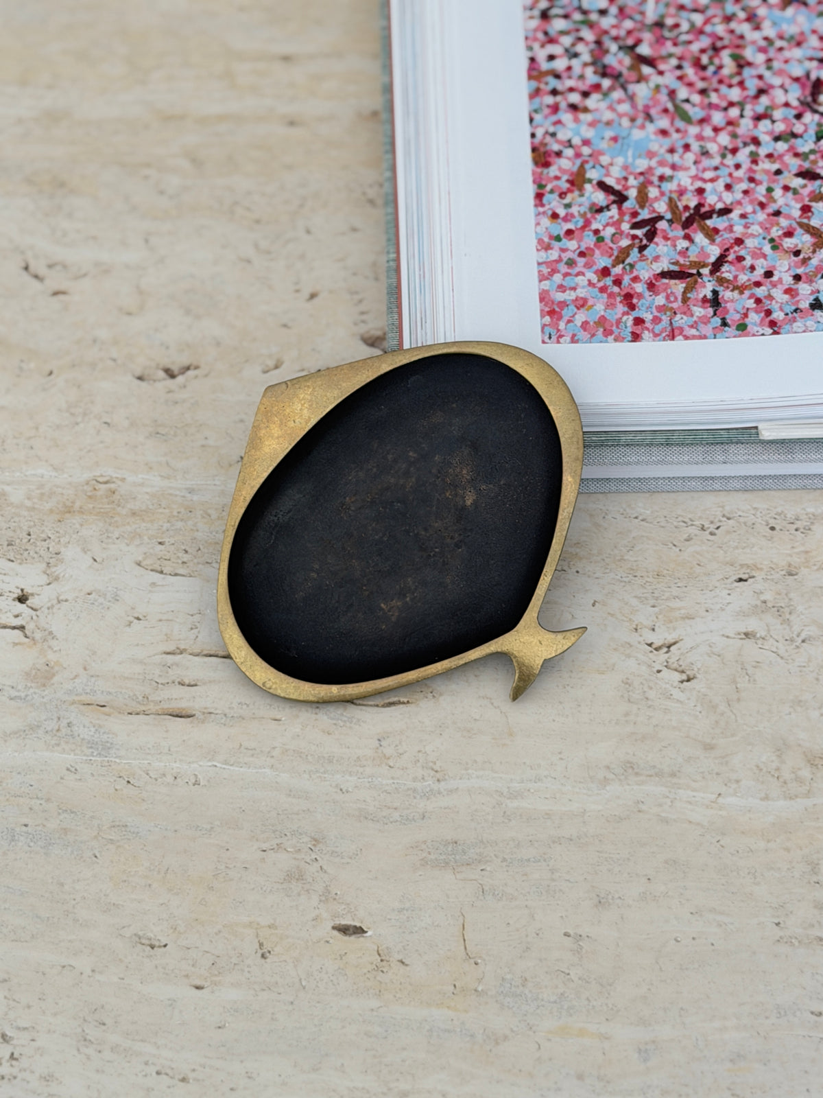 Stylized brass fish ashtray by Karl Hagenauer set against the corner of an open art book on travertine, showing its forked tail and wide brass rim