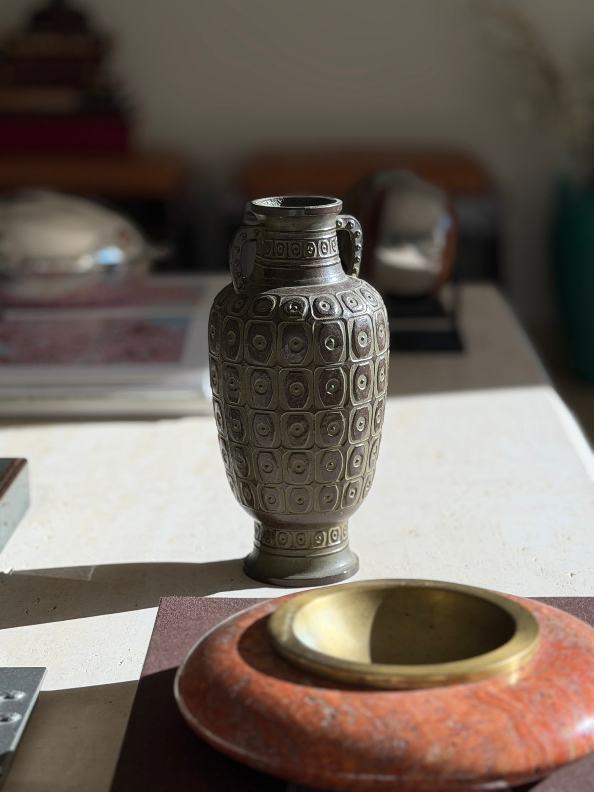 Japanese bronze vase in soft focus behind a red stone and brass ashtray, the flared foot and ring pattern band at the base