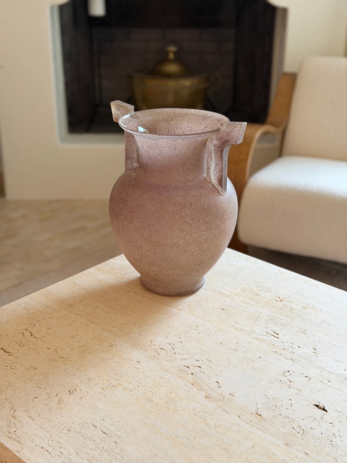 Front view of the Murano scavo glass amphora vase on a travertine table, with a brick fireplace and lidded brass pot visible behind it