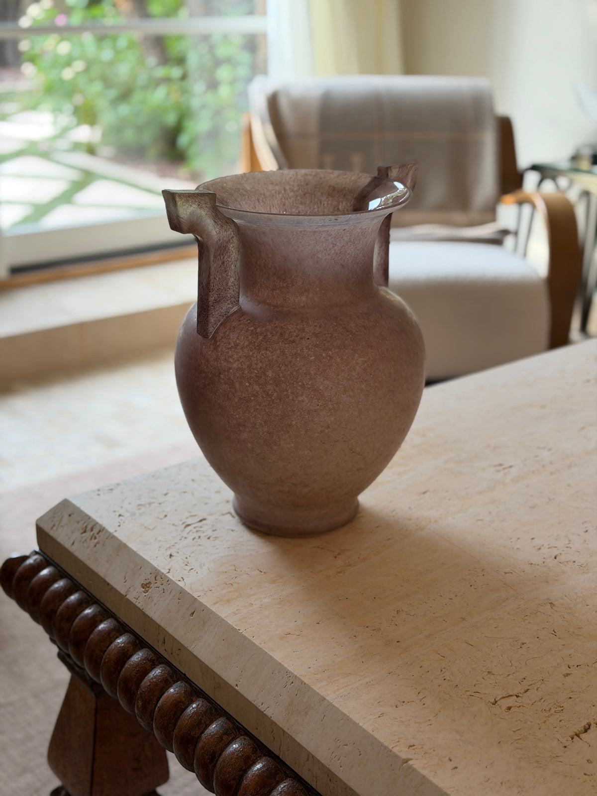 Full view of the vase at a slight angle showing its handled silhouette, with a teal ceramic vase and dried branches visible behind it