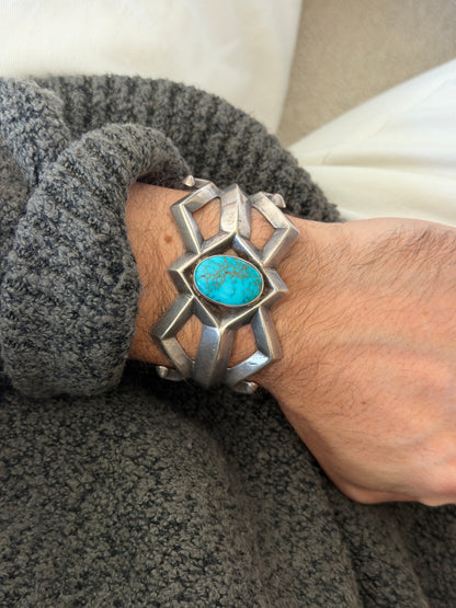 Dine sand cast silver cuff with an oval spiderweb turquoise cabochon at the center of angular open bars, worn on the wrist against a gray knit sleeve