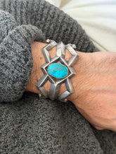 Sand cast silver cuff on the wrist, the faceted bars radiating from the bezel set turquoise toward pointed and curled terminals