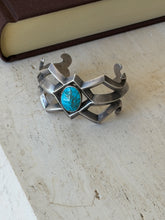 Sand cast silver and turquoise cuff resting on travertine beside a brown book, showing the full open framework and curled ends