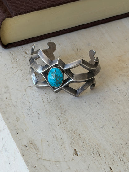 Sand cast silver and turquoise cuff resting on travertine beside a brown book, showing the full open framework and curled ends