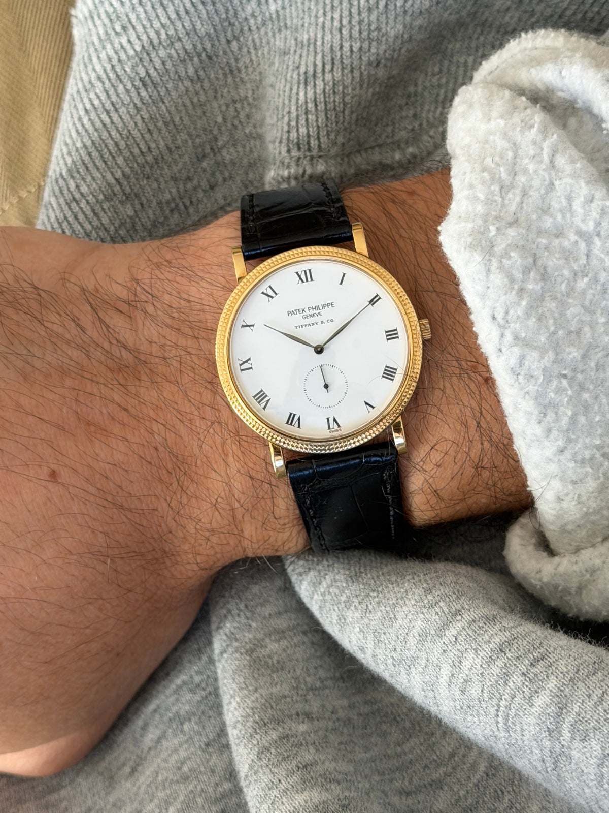Patek Philippe Calatrava worn on the wrist, close overhead view showing the hobnail bezel and black alligator strap against a gray sweater cuff