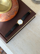 Watch laid flat on a brown book, gold buckle visible on the strap, with an orange stone bowl in the corner of the frame