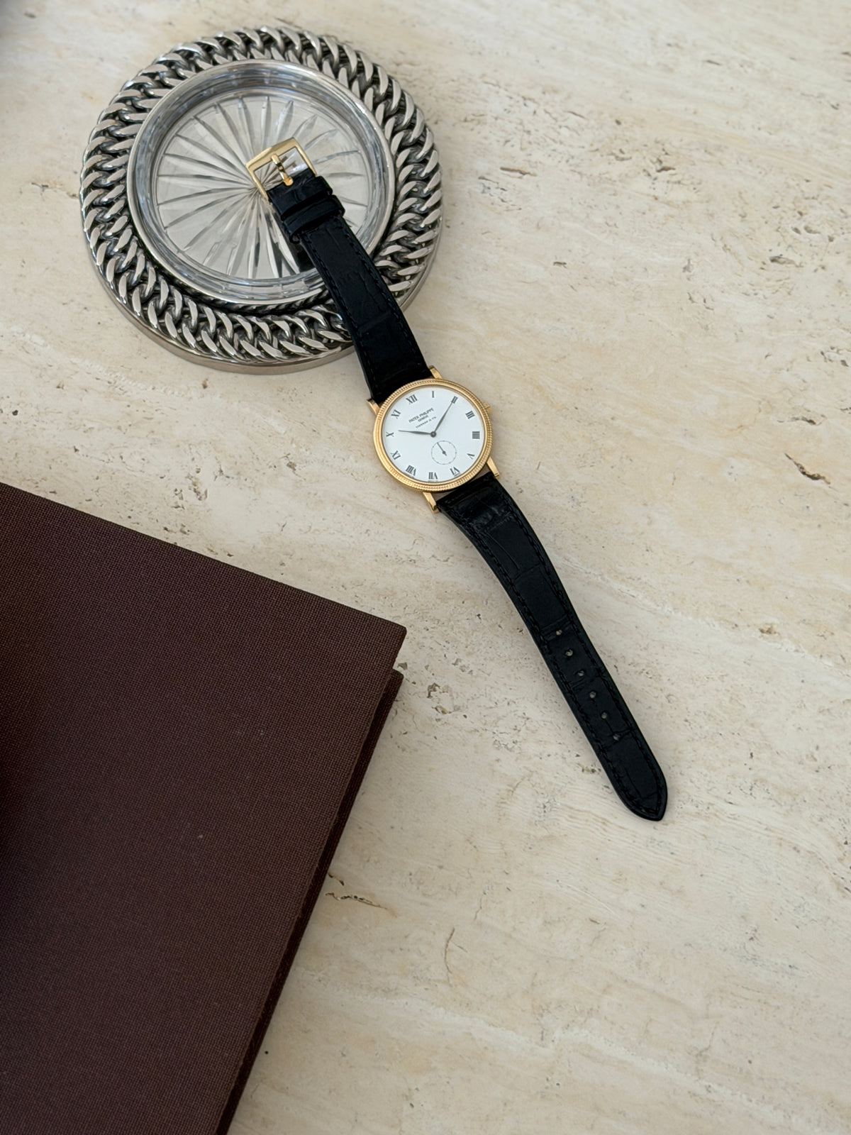 Watch resting on a stone surface beside a silver woven rim coaster, strap extended diagonally with a brown book corner in view