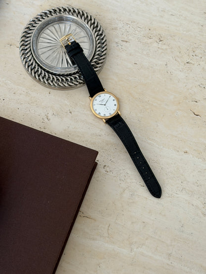 Watch resting on a stone surface beside a silver woven rim coaster, strap extended diagonally with a brown book corner in view
