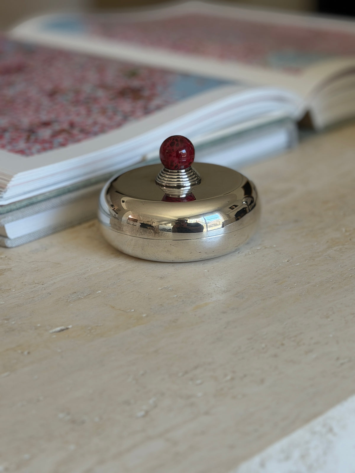 Overhead view of the Puiforcat candy dish on a travertine table near an open art book, the red finial centered on the lid