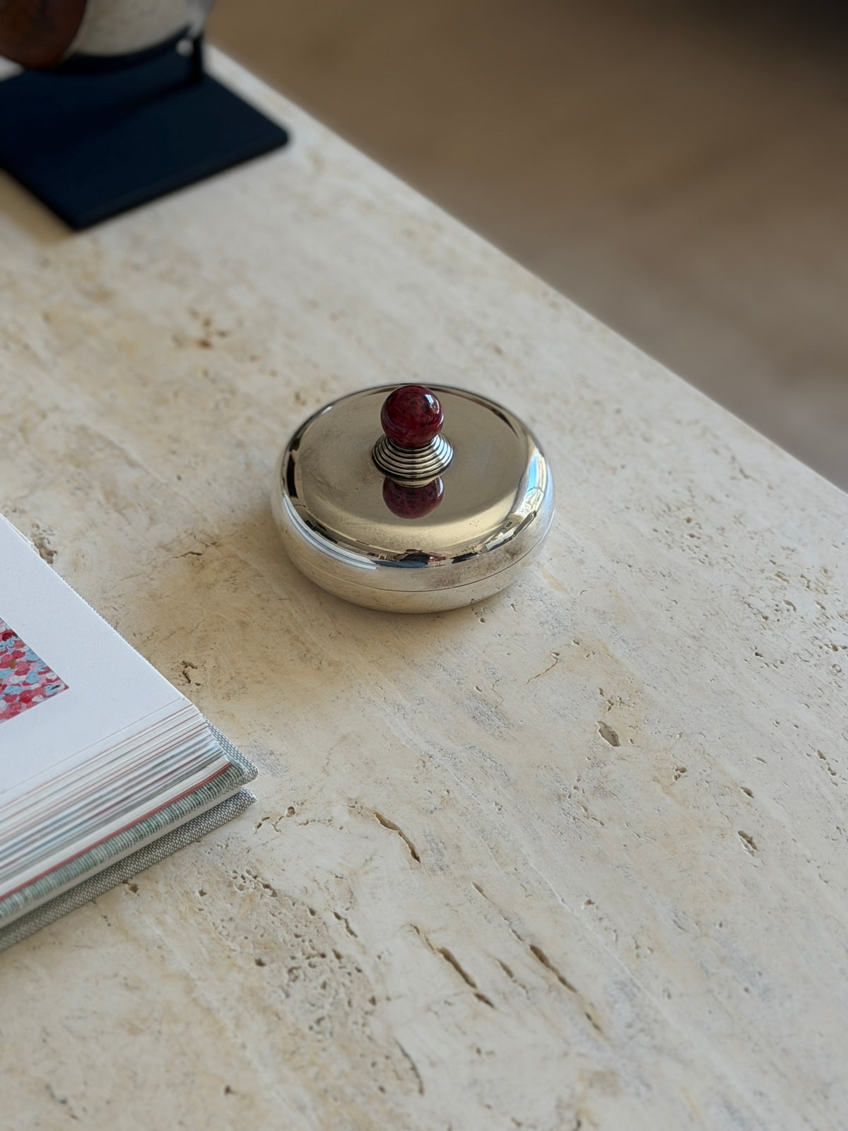 Puiforcat silver plate box in front of an open book of floral paintings, the stepped ribbed collar beneath the red stone ball