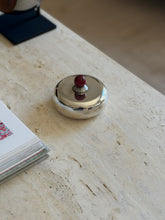 Puiforcat silver plate box in front of an open book of floral paintings, the stepped ribbed collar beneath the red stone ball